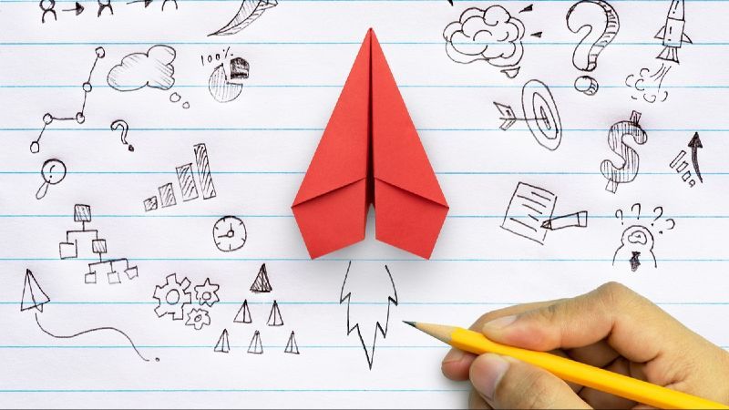 Ein roter Papierflieger liegt auf einem Blatt Papier. Ein Hand zeichnet mit einem Stift Raketenfeuer unter den Flieger, sowie Konzeptionen für ein Start-up.