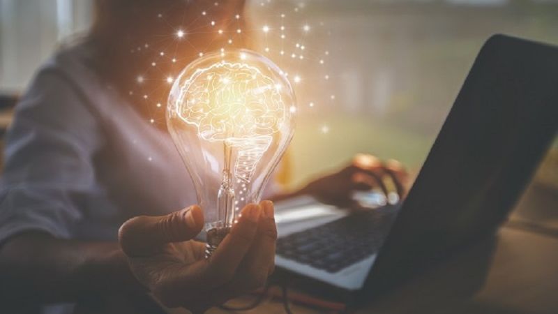 Ein Mann arbeitet am Laptop und hält gleichzeitig eine leuchtende Glühbirne, in welchem das Licht durch ein Gehirn ausgetauscht ist.