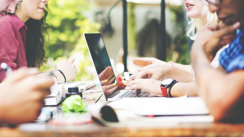 Zwei Personen mit einem Laptop sitzen weiteren Personen gegenüber